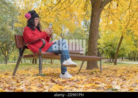 giovane donna in un cappello di lana seduta su una panchina di legno in un parco ridendo mentre chiacchiera con il suo cellulare. sfondo di alberi colorati e foglie cadute Foto Stock