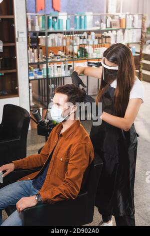stylist in visiera protettiva e guanti in lattice che tagliano i capelli di il cliente indossa una maschera medica Foto Stock