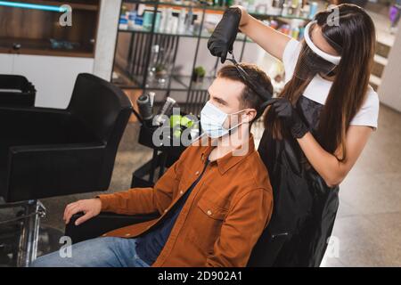 barbiere in visiera protettiva e guanti in lattice per tagliare i capelli di il cliente indossa una maschera medica Foto Stock