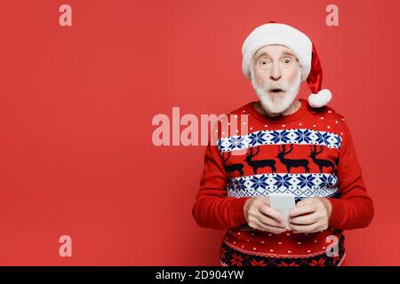 Uomo anziano scioccato in santa Hat che tiene lo smartphone isolato rosso Foto Stock