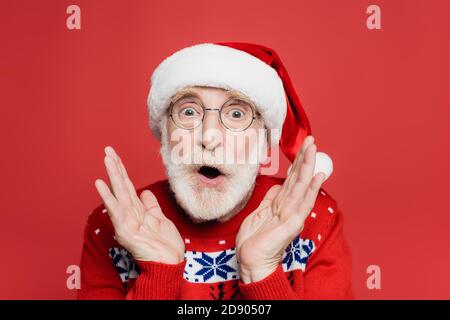 Uomo anziano scioccato nel cappello di santa e maglia che guarda sulla telecamera isolata in rosso Foto Stock