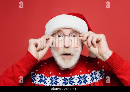 Uomo anziano scioccato in cappello di santa e occhiali che guardano telecamera isolata in rosso Foto Stock