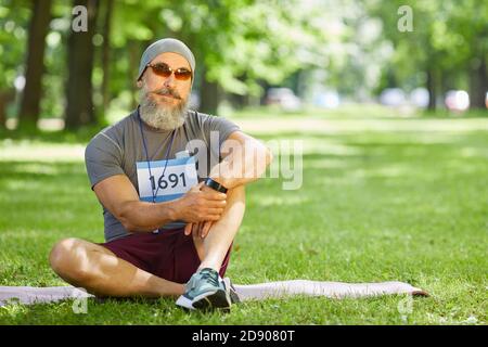 Ritratto dell'elegante uomo anziano con barba sulla presa del viso parte nella maratona estiva seduta rilassata sull'erba Foto Stock