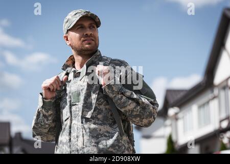Uomo di servizio militare adulto in camouflage guardando via con case offuscate sullo sfondo Foto Stock