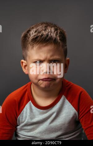 Il ragazzino è naughty e grimaces con disappunto. Il bambino scontento mostra le sue emozioni. Studio ritratto su sfondo grigio Foto Stock