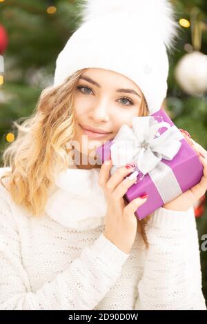 Bella ragazza in pullover bianco e cappuccio tiene un regalo di Natale ben imballato sorridente. Foto Stock