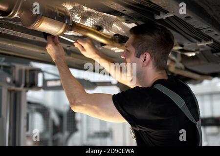 giovane meccanico di automobile maschio in automobile di controllo uniforme in servizio di automobile con veicolo sollevato, bel ragazzo che lavora duro sotto condizione di automobile sul concetto di riparazione di automobile di lifter. automobilistico Foto Stock
