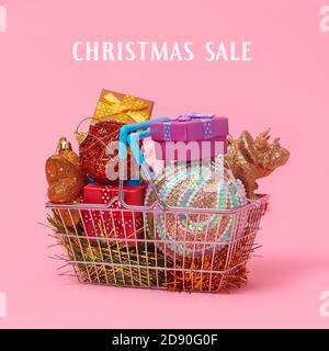 un cestino della spesa pieno di ornamenti di natale e piccole scatole del regalo, e il testo di vendite di natale su uno sfondo rosa Foto Stock