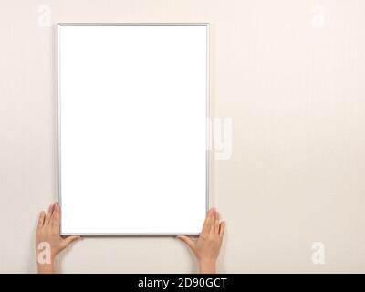 Cornice bianca in argento appesa su una parete beige. Le mani tengono il mockup della cornice fotografica sulla parete. Cornice bianca mockup Foto Stock