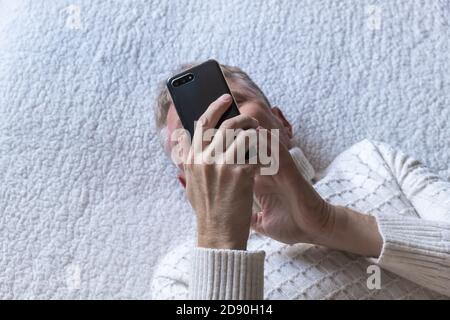 Un uomo di mezza età si trova sul divano e guarda le notizie sul suo cellulare. Messaggi, controllo del mercato azionario. Messa a fuoco selettiva.primo piano. Vista dall'alto Foto Stock