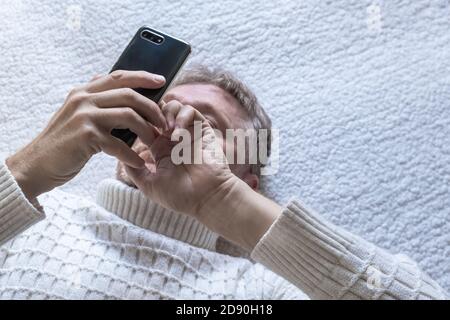 Un uomo di mezza età si trova sul divano e guarda le notizie sul suo cellulare. Messaggi, controllo del mercato azionario. Messa a fuoco selettiva.primo piano. Vista dall'alto Foto Stock