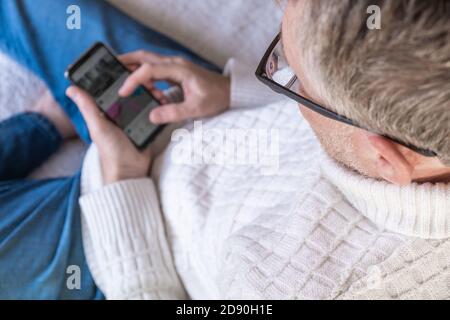 Un uomo di mezza età si siede sul divano e guarda le notizie sul suo cellulare. Controllo del mercato azionario, acquisti online, messaggistica. Messa a fuoco selettiva. Foto Stock