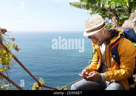 Ritratto di felice uomo di viaggio afroamericano seduto in mare guardando il telefono cellulare Foto Stock