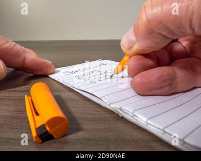 Primo piano POV di una mano di un uomo che tiene una penna di feltro arancione e che scrive una lista di negozi di generi alimentari su un blocco note allineato su una scrivania. Foto Stock