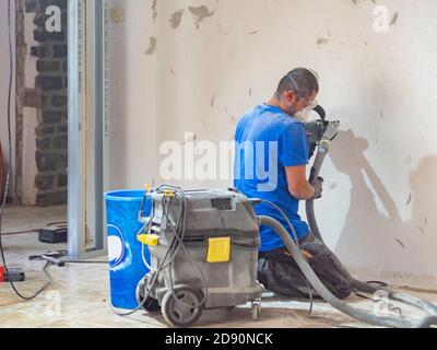 il lavoratore leviga la parete con un macchinario con maschera Foto Stock