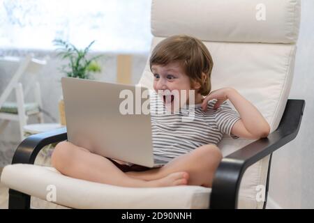 Felice ragazzo sorpreso sedersi sul computer utilizzando il computer portatile e fare una videochiamata. Il bambino sorridente seduto a casa riposa sulla sedia occupata da gadget. Online Foto Stock