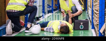 Il lavoratore del magazzino panoramico fa il primo soccorso al suo collega che si trova sul pavimento del magazzino dopo l'incidente mentre lavora. Primo utilizzo per la sicurezza industriale Foto Stock