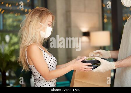 adorabile cliente donna prendere il suo ordine in ristorante durante il coronavirus, cameriere personale di caffè dare cibo in contenitore indossando guanti di protezione, la donna è in maschera medica Foto Stock