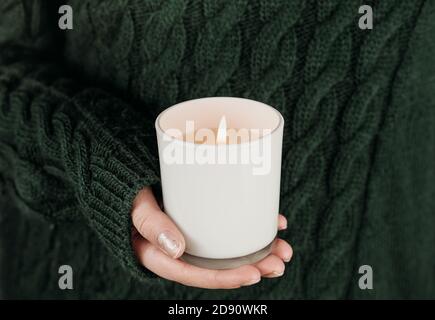 donna mano che tiene la candela bianca di soia. ragazza adolescente che indossa il maglione verde e che tiene la candela bruciante. concetto di festa. esperienza spirituale e pace interiore. fuoco selettivo Foto Stock