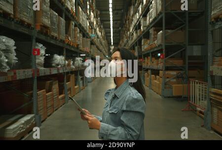 Donna che lavora in Asia indossa una maschera utilizzando il controllo digitale del tablet scorte di prodotti in magazzino Foto Stock