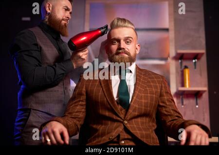 elegante uomo seduto in barbiere mentre il parrucchiere asciuga i capelli, bello soddisfatto giovane bearded ragazzo caucasico ottenere trendy taglio di capelli, barbiere servendo cliente Foto Stock