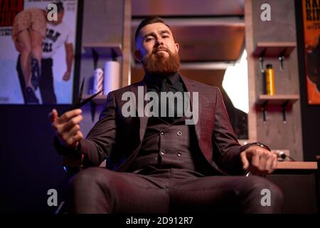 barbiere forbici, barbiere, uomo brutale con forbici, uomo elegante, seduto al suo posto di lavoro in camera oscura Foto Stock