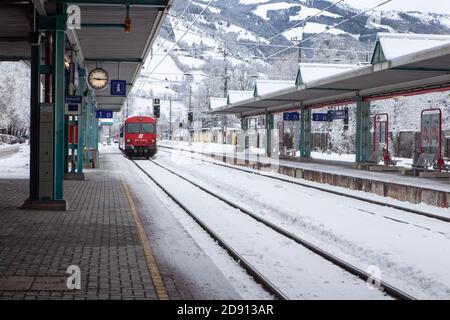 Austria / Zell am See - Febbraio 06 2013: Il treno ad alta velocità per passeggeri rossi si ferma alla stazione ferroviaria deserta coperta di neve in montagna. Conce Foto Stock