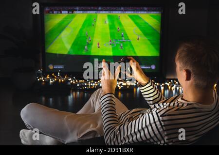 Giovane uomo hipster che gioca a video gioco di calcio calcio su console. Giocatore con controller gamepad tenere il joystick wireless seduto sul pouf di notte. Foto Stock