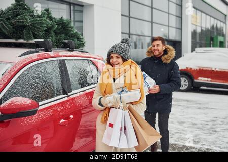 La giovane coppia con le scatole del regalo è vicino automobile con l'albero sulla parte superiore. Insieme all'aperto in inverno Foto Stock