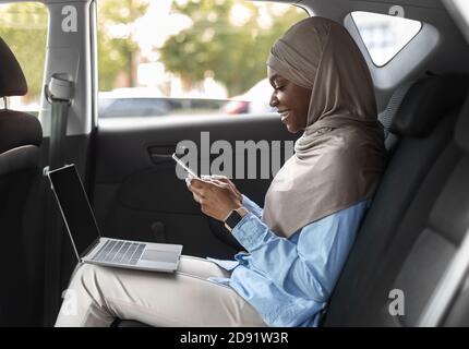 Businesswoman musulmano africano occupato usando il laptop e lo smartphone mentre Equitazione Auto Foto Stock