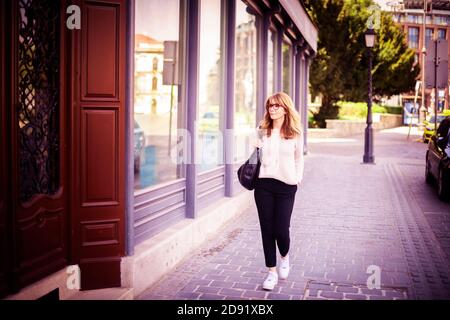 Foto di sorridente donna di mezza età che indossa casual elegante mentre si cammina da sola sulla strada in centro. Foto Stock