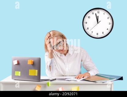 Donna di affari matura che si sente depressa sul posto di lavoro, manca la scadenza, non riuscendo a gestire il suo tempo su sfondo blu Foto Stock