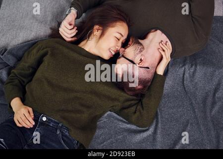 Felice coppia multietnica in abiti casual sdraiati insieme all'interno dello studio. Ragazzo caucasico con ragazza asiatica Foto Stock