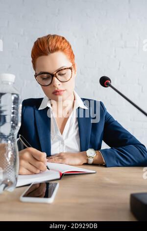 Donna rossa in abbigliamento formale, scrittura in taccuino, mentre si siede al tavolo con bottiglia d'acqua e microfono Foto Stock