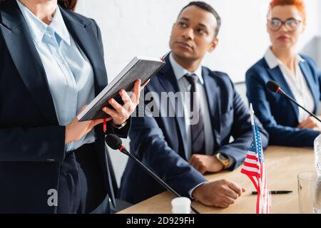 I membri del partito politico che guardano il collega in piedi con il taccuino dentro sala riunioni su sfondo sfocato Foto Stock