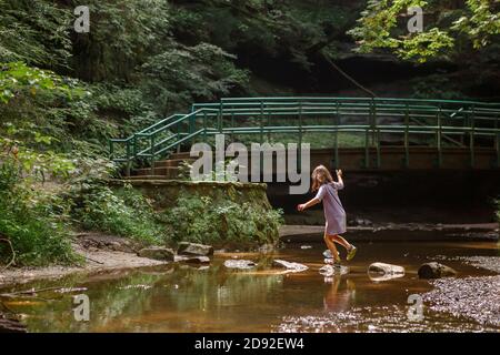 Una ragazza piccola equilibra sulle pietre di passo attraverso un ruscello in legno Foto Stock