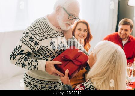 Donna anziana felice che prende la scatola del regalo dal marito, seduto con la famiglia a casa Foto Stock