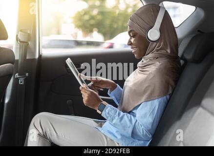 Intrattenimento in auto. Donna musulmana nera che usa la tavoletta digitale mentre guida l'automobile Foto Stock