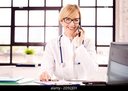 Portrat ha girato di sorridente dottoressa seduto alla scrivania e di fare una chiamata con la paziente mentre lavora in ufficio medico. Foto Stock