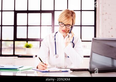 Portrat ha girato di sorridente dottoressa seduto alla scrivania e di fare una chiamata con la paziente mentre lavora in ufficio medico. Foto Stock