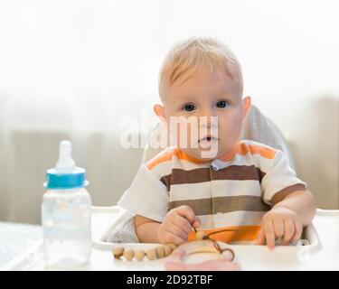 Un piccolo bambino sorpreso dall'occhio blu si siede a un tavolo e guarda la fotocamera. Sul tavolo c'è un biberon con un succhietto e acqua. Stupore Foto Stock