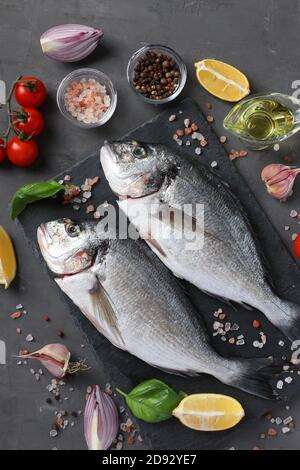 Due pesci crudi dorado con ingredienti e condimenti come basilico, limone, sale, pepe, pomodori ciliegini e olio d'oliva su sfondo scuro, vista dall'alto Foto Stock