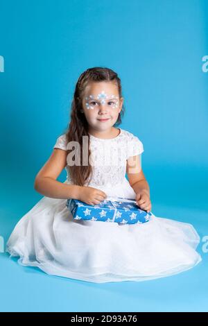 Una ragazza in un vestito bianco e con il trucco di Natale sulla sua faccia che tiene un regalo blu isolato su un sfondo azzurro Foto Stock