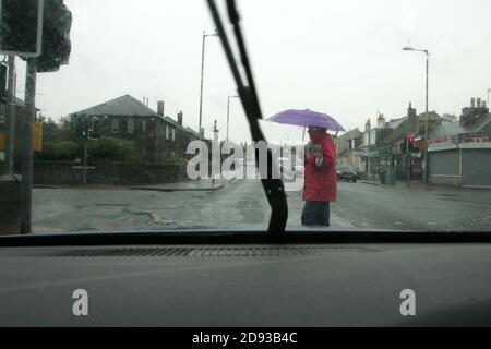 Ayr, Ayrshire, Scozia , Regno Unito . Una giacca rossa donna sotto la pioggia che tiene un ombrello attraversa di fronte all'auto su un passaggio pedone pellicano Foto Stock