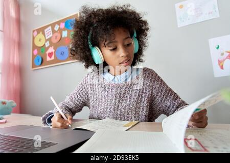 La ragazza africana intelligente del capretto indossa le cuffie che imparano in linea facendo i compiti a casa. Foto Stock