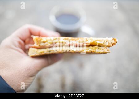 Tenere a mano un panino di formaggio e prosciutto di fronte una tazza di caffè sfocata Foto Stock