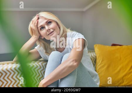 Donna caucasica di media età che sogna mentre si siede con la testa rialzata su un comodo divano nel suo appartamento, visto attraverso la pianta verde. Persone che trascorrono il tempo a casa Foto Stock