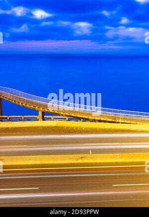 Lunga esposizione di sentieri di luce dal traffico sulla strada costiera con passerella e oceano in background all'alba. Foto Stock