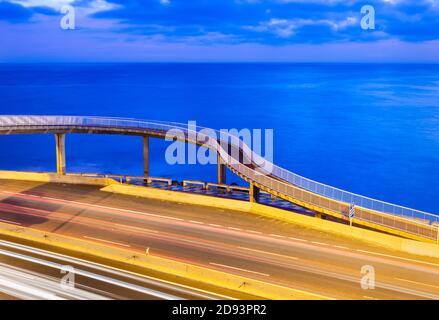 Lunga esposizione di sentieri di luce dal traffico sulla strada costiera con passerella e oceano in background all'alba. Foto Stock