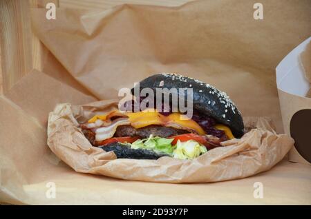 Hamburger di manzo con un panino nero, con lattuga e maionese e ketchup servito su pezzi di carta marrone su un rustico tavolo di legno di bancone, al buio Foto Stock